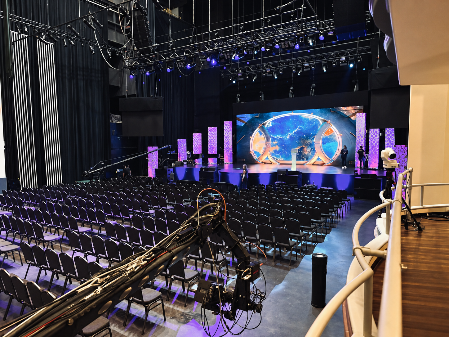 Full theatre view showing the stage, LED screen, rows of seats, overhead lighting, and a production camera crane