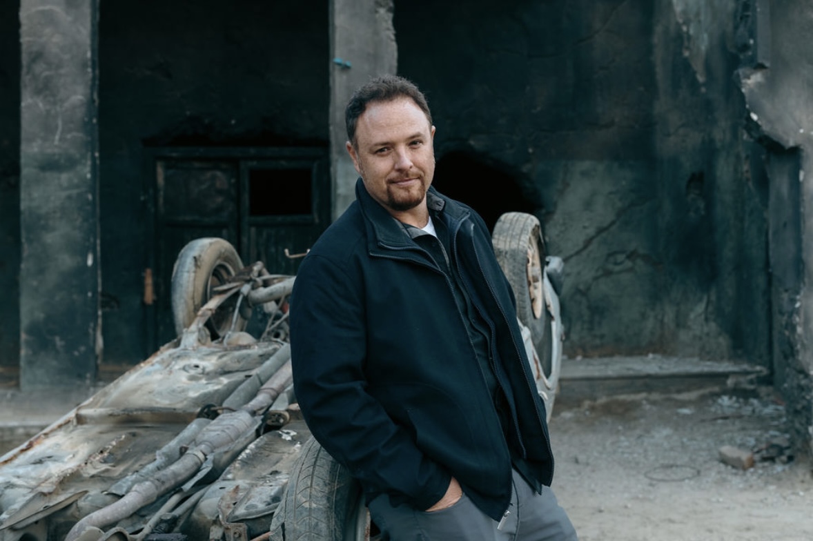 Wayne standing beside a wooden cart on a period film set